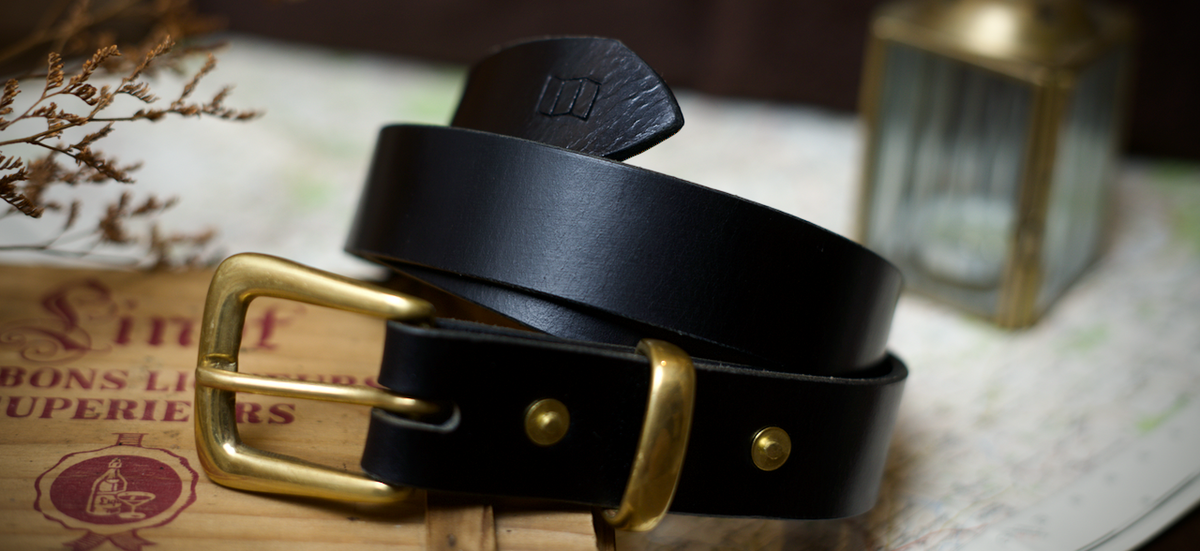 A black leather full grain handmade belt with a brass buckle, coiled on a wooden surface with vintage map and dried floral props in the background.